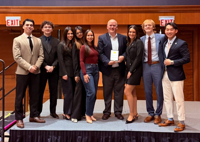 Nine people, dressed in business and semi-formal attire, stand together on a stage; a man in the center holds an American Marketing Association award plaque.