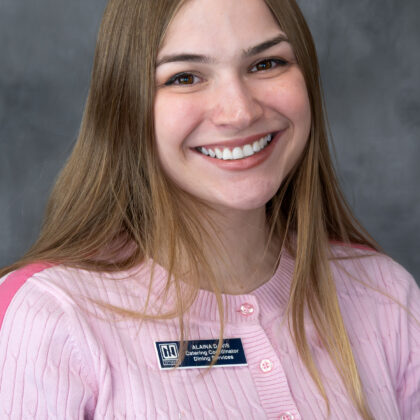 A woman with long blonde hair, wearing a light pink sweater and a name badge, smiles at the camera against a gray background.