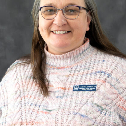 A woman with long brown hair and glasses, wearing a pink patterned sweater and a name badge, smiles in front of a gray background.