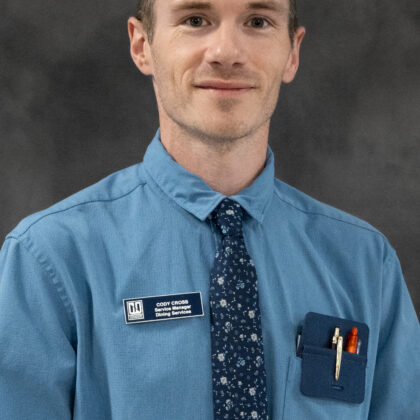 A man in a blue shirt and patterned tie stands against a gray background, wearing a name badge and pens in his shirt pocket.