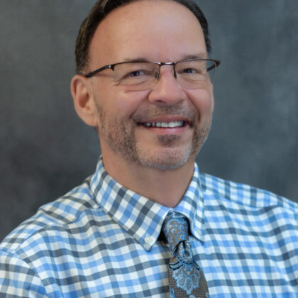A man with glasses, short hair, and a beard, wearing a blue checked shirt, paisley tie, and a name badge, poses in front of a plain gray background.
