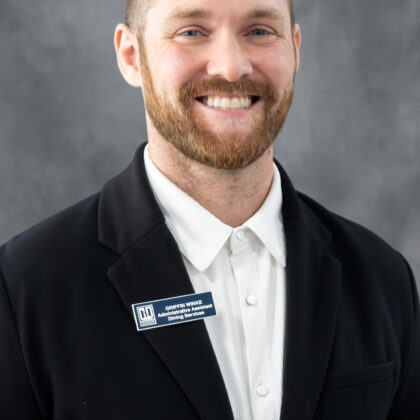 A man wearing a black suit jacket and a white shirt stands smiling in front of a gray background, with a name badge on his lapel.