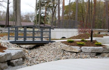 A landscaped garden on the Northwood University campus walk features two small arched bridges over a dry rock creek, surrounded by trees, large stones, and mulched planting beds—a peaceful space for community gatherings.