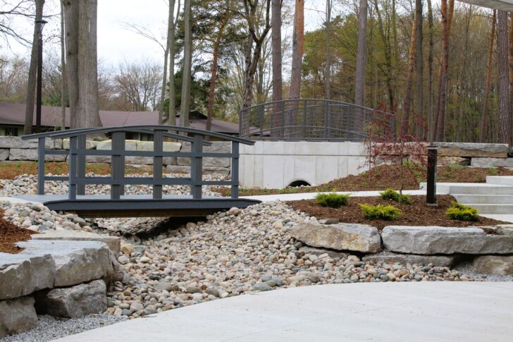 A landscaped garden on the Northwood University campus walk features two small arched bridges over a dry rock creek, surrounded by trees, large stones, and mulched planting beds—a peaceful space for community gatherings.