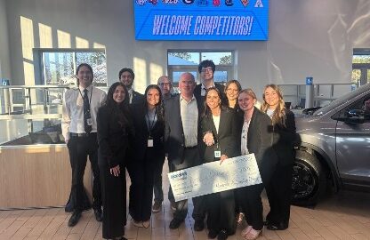 A group of people in business attire pose indoors, holding a large check, with a "Welcome Competitors!" sign and university logos displayed on a screen in the background.