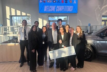 A group of people in business attire pose indoors, holding a large check, with a "Welcome Competitors!" sign and university logos displayed on a screen in the background.