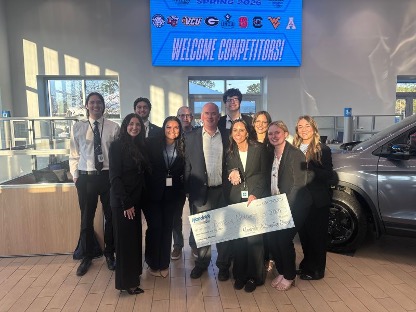 A group of people in business attire pose indoors, holding a large check, with a "Welcome Competitors!" sign and university logos displayed on a screen in the background.