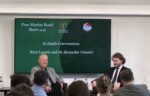Two men sit on a stage in front of an audience, speaking during an event titled "Free Market Road Show 2026" with the Prosperity Institute logo displayed on a screen behind them.