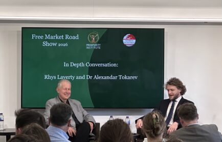 Two men sit on a stage in front of an audience, speaking during an event titled "Free Market Road Show 2026" with the Prosperity Institute logo displayed on a screen behind them.