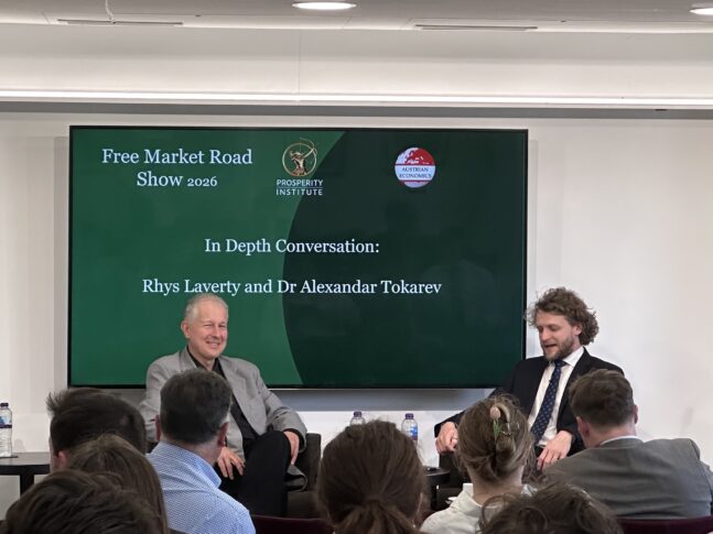 Two men sit on a stage in front of an audience, speaking during an event titled "Free Market Road Show 2026" with the Prosperity Institute logo displayed on a screen behind them.