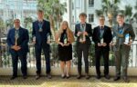 Six people in formal attire stand in a row indoors, each holding trophies and wearing medals, with palm trees and large windows in the background.