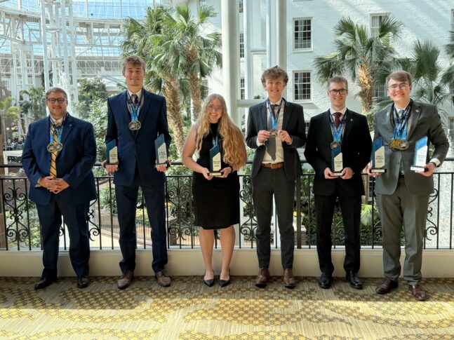 Six people in formal attire stand in a row indoors, each holding trophies and wearing medals, with palm trees and large windows in the background.