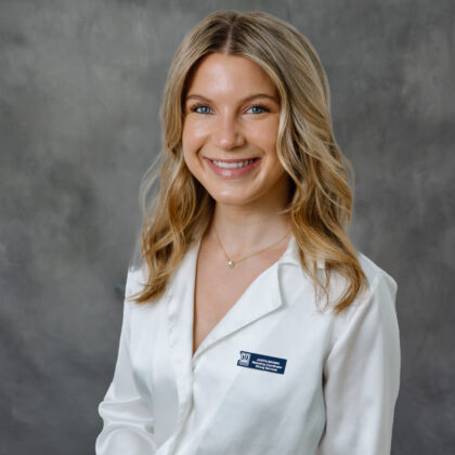 A woman with long blonde hair wearing a white blouse and name badge, smiling in front of a gray studio backdrop.