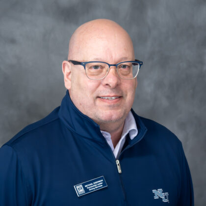 A man with glasses wearing a dark blue pullover with an NSU logo and a name badge poses in front of a gray background.