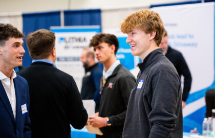 Several young men in business attire network at an indoor career fair, featuring informational booths and banners, including those representing top sales programs like Northwood University, recognized nationally.