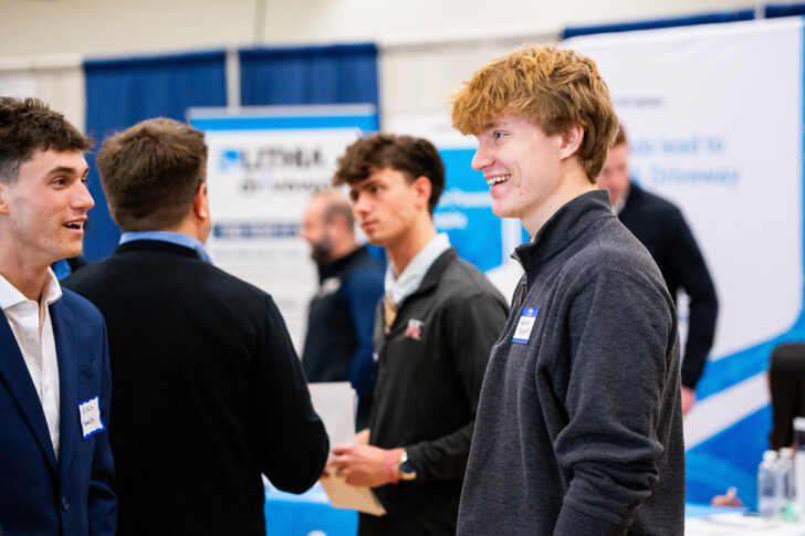 Several young men in business attire network at an indoor career fair, featuring informational booths and banners, including those representing top sales programs like Northwood University, recognized nationally.