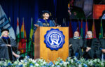 A woman in academic regalia speaks at a podium during a university graduation ceremony, with officials seated behind her and international flags displayed in the background.