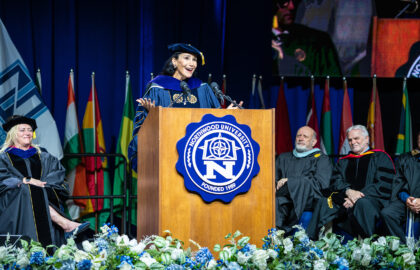 A woman in academic regalia speaks at a podium during a university graduation ceremony, with officials seated behind her and international flags displayed in the background.