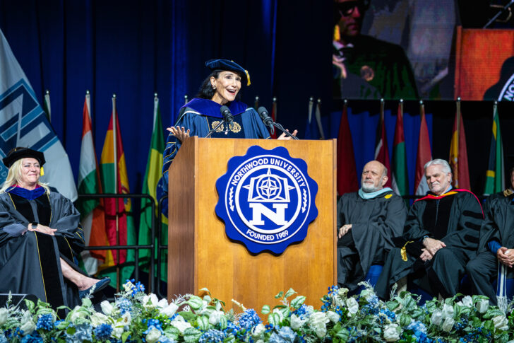 A woman in academic regalia speaks at a podium during a university graduation ceremony, with officials seated behind her and international flags displayed in the background.