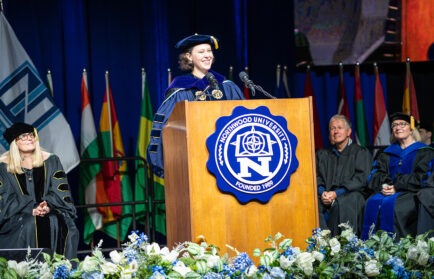 A person in academic regalia speaks at a podium with the Northwood University emblem, addressing the Class of 2026, as officials and international flags stand behind them. The spirit of freedom fills the stage.