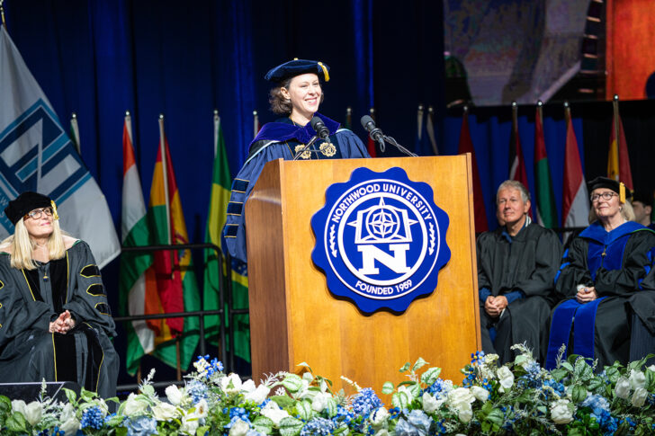 A person in academic regalia speaks at a podium with the Northwood University emblem, addressing the Class of 2026, as officials and international flags stand behind them. The spirit of freedom fills the stage.