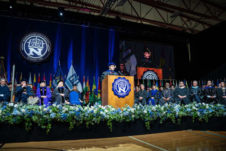 A speaker in academic regalia addresses an audience from a podium at a university commencement celebration, with faculty and officials seated on stage, highlighting the balance of freedom and responsibility that accompanies graduation.