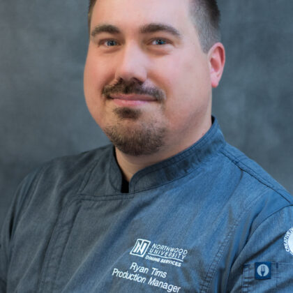 A man in a blue chef’s jacket with "Production Manager" embroidered on it poses for a professional portrait against a gray background.
