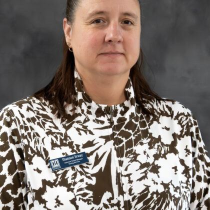 A woman with brown hair pulled back, wearing a white and brown patterned top and a name tag that reads "Shannon Envy," stands in front of a gray backdrop.