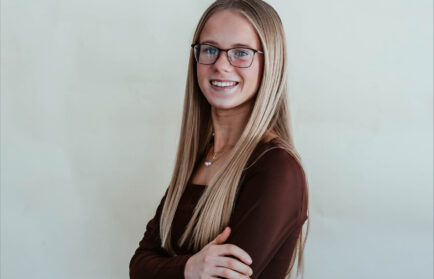 A young woman with long straight blonde hair and glasses stands smiling with arms crossed, proudly representing the Mallorie Hollenbeck NIADA $10,000 Scholarship, wearing a brown long-sleeve top against a plain light background.