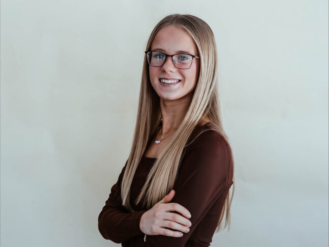 A young woman with long straight blonde hair and glasses stands smiling with arms crossed, proudly representing the Mallorie Hollenbeck NIADA $10,000 Scholarship, wearing a brown long-sleeve top against a plain light background.