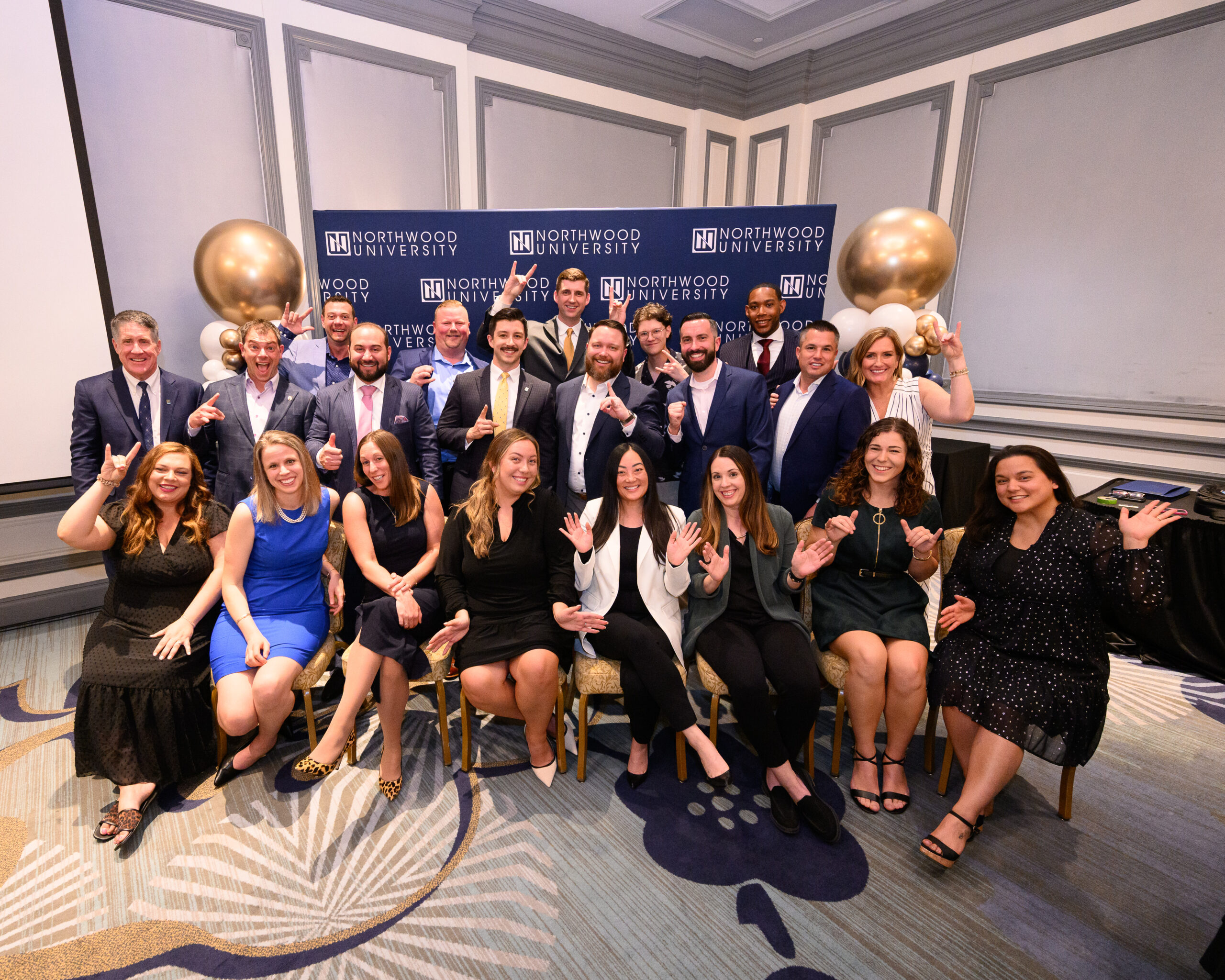 A group of people pose together indoors in front of a Northwood University banner, some seated and some standing, celebrating Northwood's Project 100 with gold and white balloons in the background.