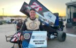 A smiling race car driver in a suit holds a "Northwood University Heat Race Winner" sign in front of his sprint car at a racetrack.