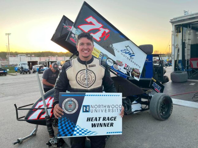 A smiling race car driver in a suit holds a "Northwood University Heat Race Winner" sign in front of his sprint car at a racetrack.