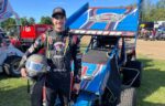 A race car driver in a racing suit stands next to a blue sprint car, holding a helmet—Charlie Baur showcases both his motorsports career and business discipline, with other vehicles and people visible in the background.