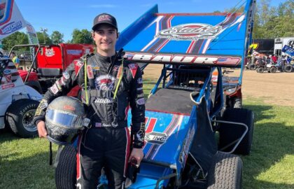 A race car driver in a racing suit stands next to a blue sprint car, holding a helmet—Charlie Baur showcases both his motorsports career and business discipline, with other vehicles and people visible in the background.