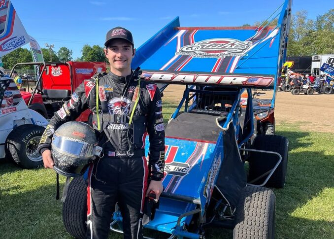 A race car driver in a racing suit stands next to a blue sprint car, holding a helmet—Charlie Baur showcases both his motorsports career and business discipline, with other vehicles and people visible in the background.
