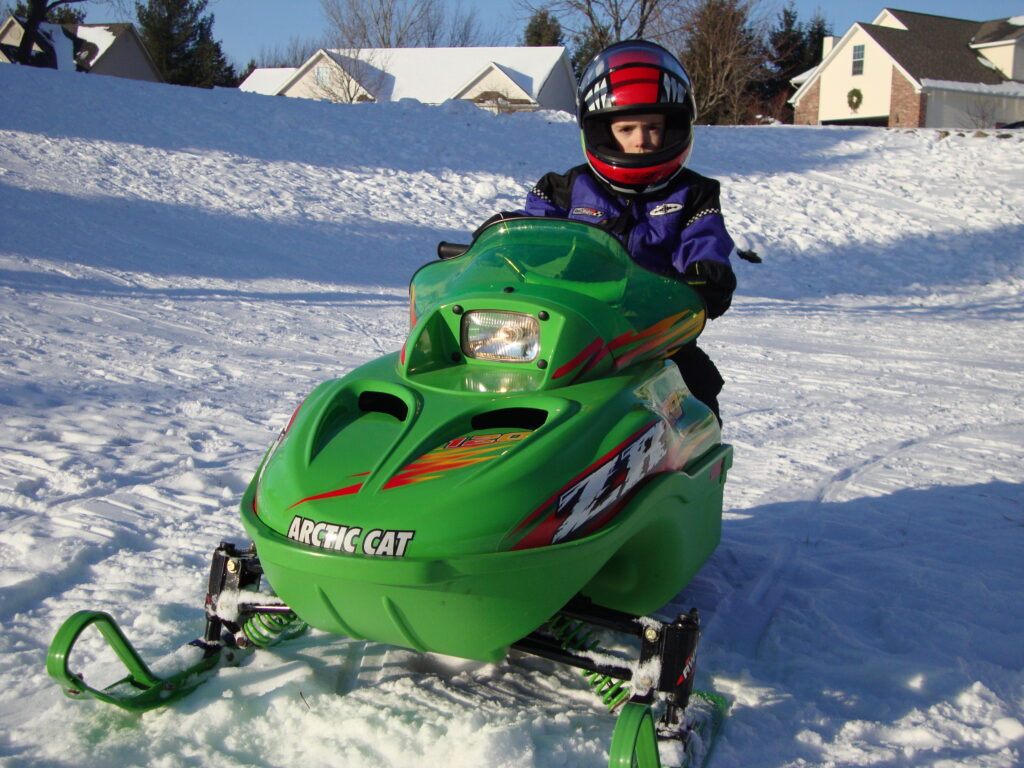 Charlie Baur, known for his motorsports career and business discipline, rides a green Arctic Cat snowmobile on a snowy surface, wearing a helmet and winter gear, with houses and trees visible in the background.
