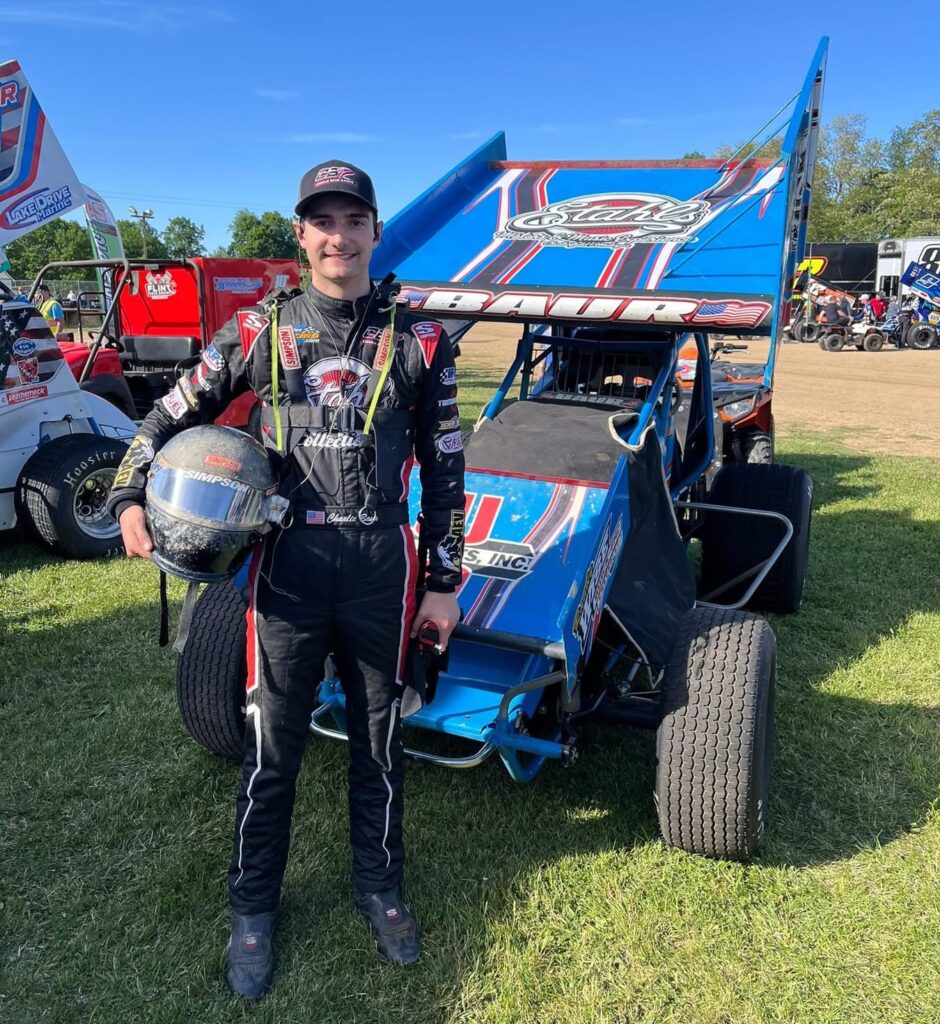 A racecar driver in full gear stands on grass, holding a helmet, in front of Charlie Baur's blue sprint car with sponsor logos—a perfect snapshot of a focused motorsports career on a sunny day.