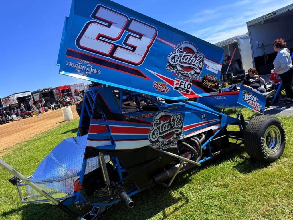 Blue and red sprint car number 23, driven by Charlie Baur, parked on grass near a dirt racetrack—trailers and people in the background reflect the lively atmosphere of a dedicated motorsports career.