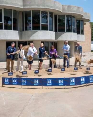 Seven people in business attire participate in a groundbreaking ceremony at Northwood University, using shovels to move dirt in front of a modern building near the Sybil Ludington statue.