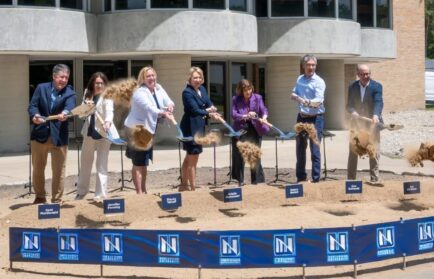 Seven people in business attire participate in a groundbreaking ceremony at Northwood University, using shovels to move dirt in front of a modern building near the Sybil Ludington statue.
