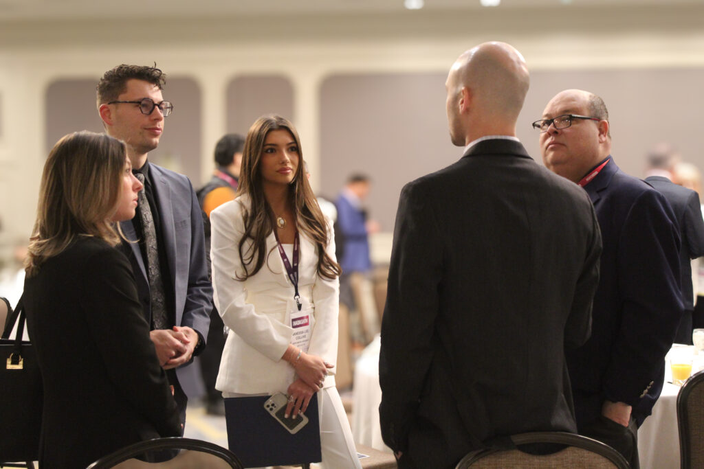Five people in business attire are standing and having a conversation in a university conference room, with tables, chairs, and automotive brochures visible in the background.
