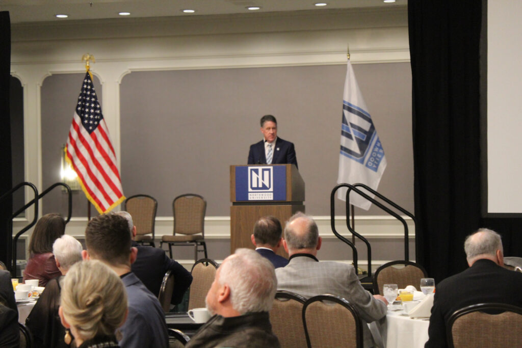 A man speaks at a podium in a banquet hall, with people seated at tables, an American flag, and a university automotive corporate flag in the background.