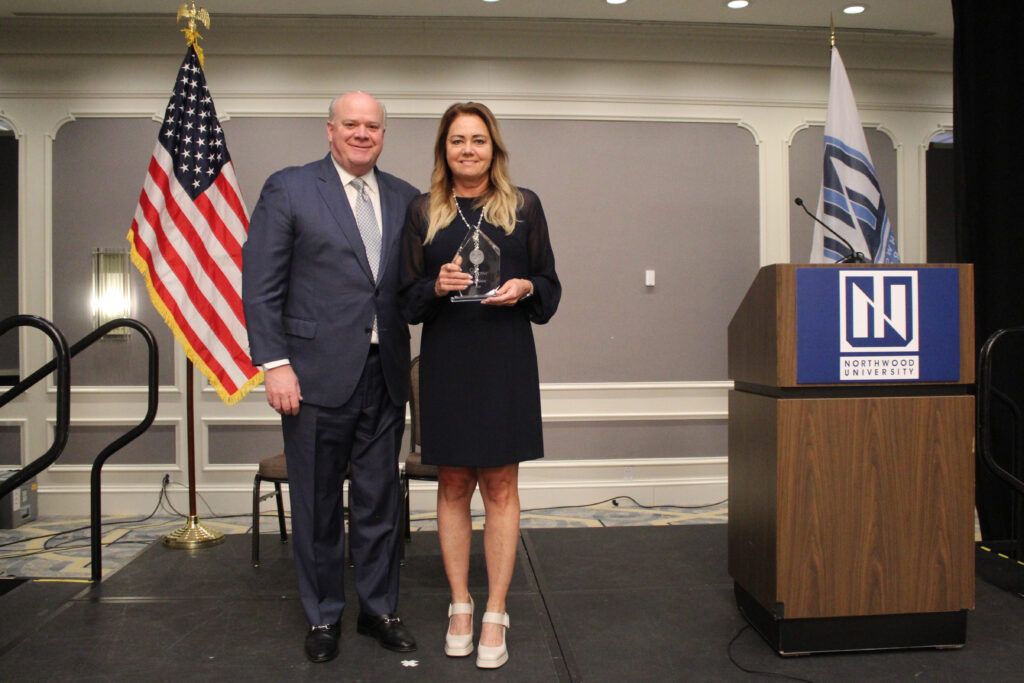 A man in a suit and a woman holding a glass award stand on stage near a podium with the Northwood University logo, surrounded by flags, at a prestigious university automotive event.