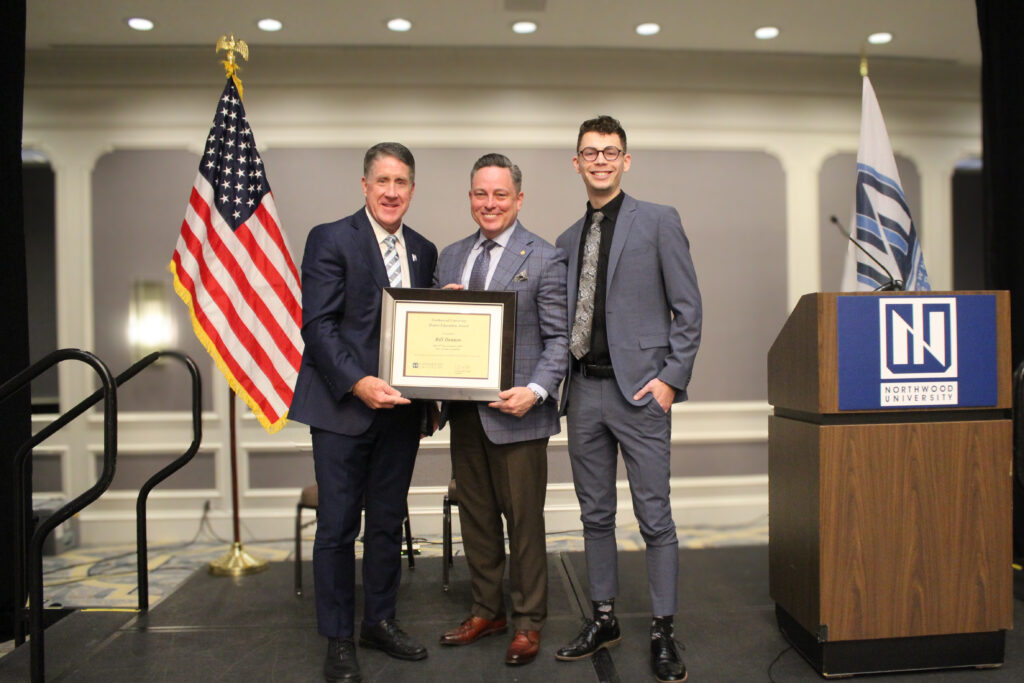 Three men stand on a stage; two hold a framed certificate. An American flag and a Northwood University automotive podium are visible in the background.