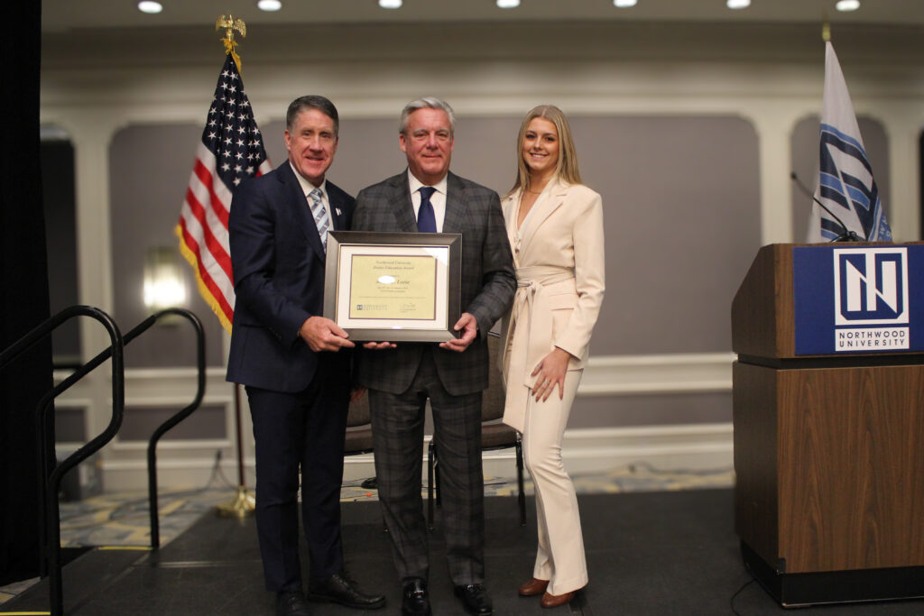 Three people stand indoors at a university event; the man in the center holds a framed certificate. An American flag and a podium with the Northwood University logo highlight the automotive-focused celebration.