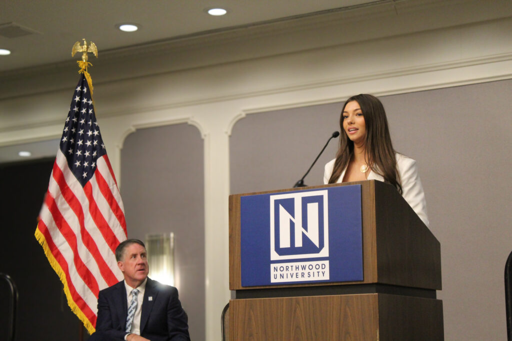 A woman speaks at a podium with a Northwood University sign, highlighting the university's automotive excellence, while a man sits nearby. An American flag stands in the background.