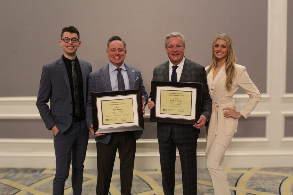 Four people pose indoors at a university event; two men in suits hold framed certificates while another man and a woman, involved in automotive research, stand beside them—all smiling at the camera.