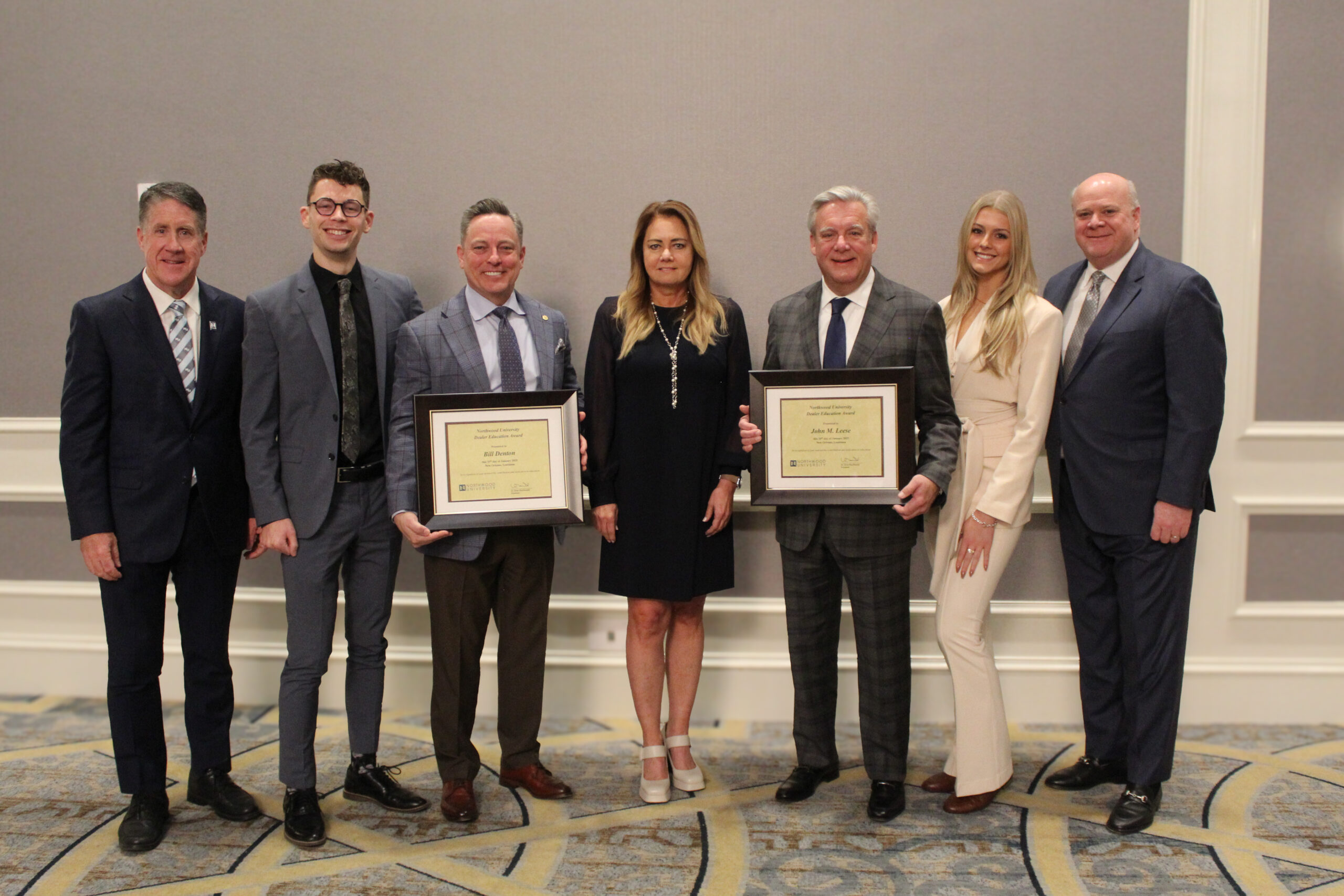 Seven people in formal attire pose in a line; two men in the center, representing university automotive excellence, are holding framed certificates.