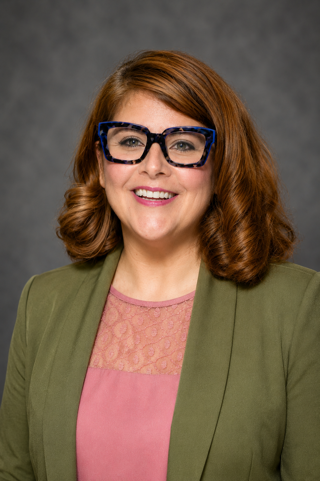 Woman with shoulder-length brown hair, wearing blue glasses, a green blazer, and a pink top, smiling in front of a gray background.